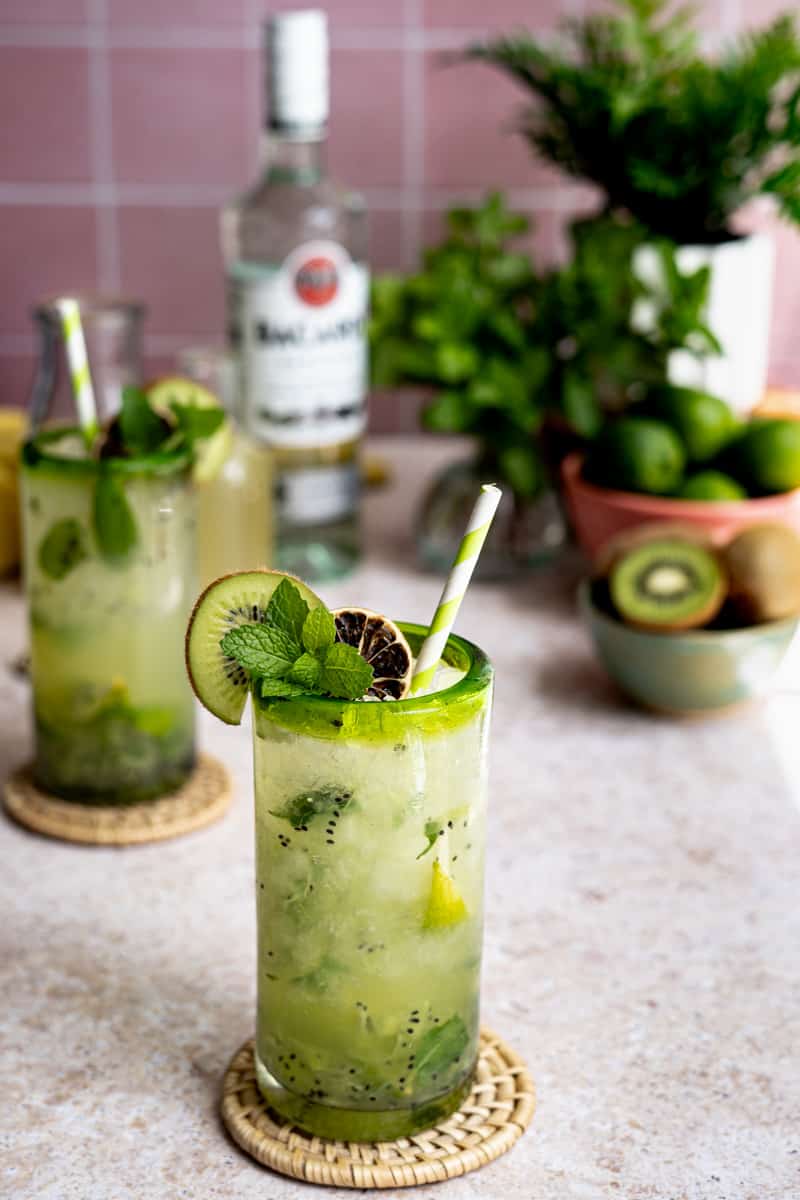 A kiwi mojito sits in the middle of a photo frame on a light brown stone countertop. Ingredients used to make the kiwi mojito are sitting in the background, along with another kiwi mojito.