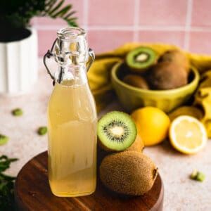 feature image of a bottle of kiwi simple syrup. A bottle of kiwi simple syrup sits on a small wooden pedestal in front of a bowl of kiwi fruits and lemon. pieces of kiwi fruit are scattered atop the counter.