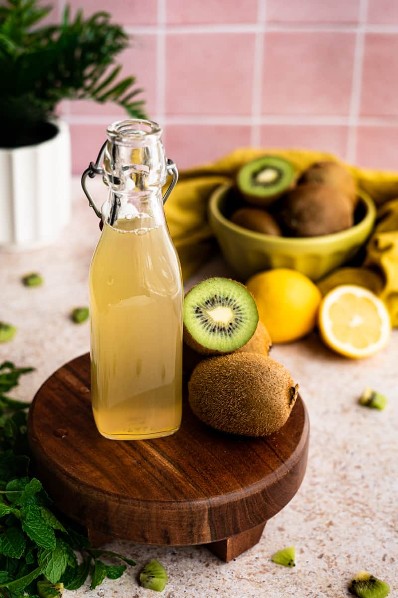a bottle of kiwi simple syrup sits on a small wooden pedestal in front of a bowl of kiwi fruits and lemon. pieces of kiwi fruit are scattered on the countertop in the foreground and backgroumd.