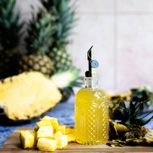 A bottle of pineapple simple syrup sits on a wooden cutting board next to a pile of pineapple chunks. There are pineapples in the background, including a sliced half of a pineapple.