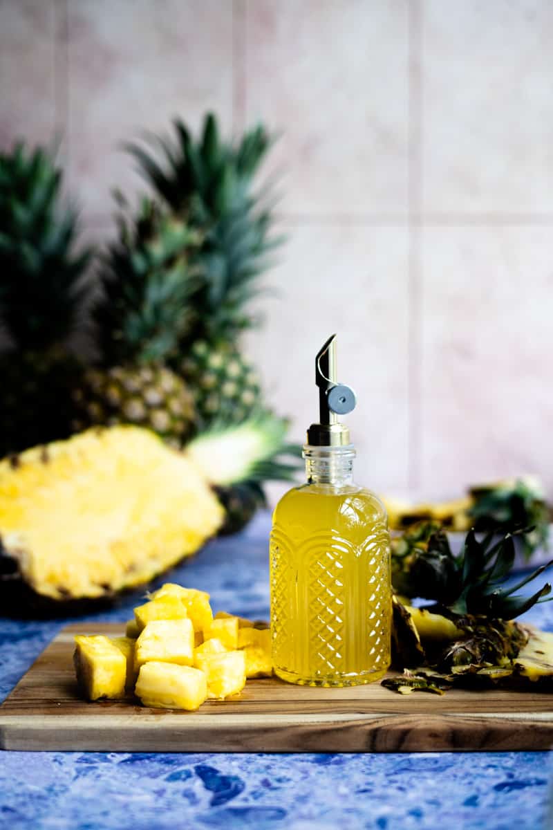 A bottle of pineapple simple syrup sits on a wooden cutting board next to a pile of pineapple chunks. There are pineapples in the background, including a sliced half of a pineapple.