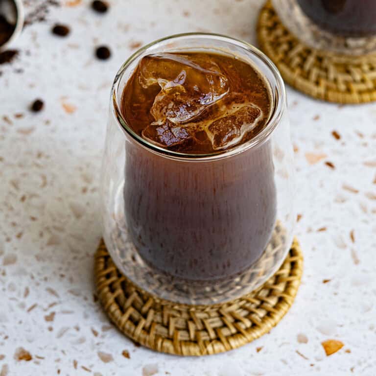 A clear glass of iced Americano coffee with large ice cubes sits on a woven coaster atop a speckled white surface. Another glass and coffee beans are partially visible in the background.