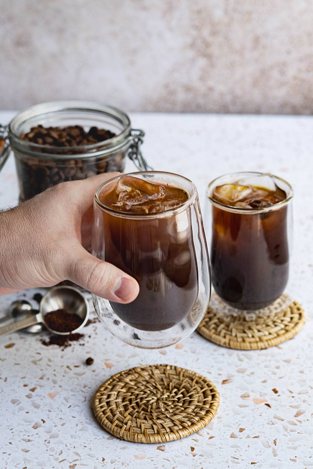 A hand holds a glass of iced americano coffee, with another glass on a woven coaster nearby. In the background, a jar of coffee beans and a metal scoop rest on a speckled surface.
