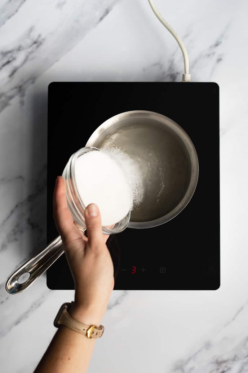 Pouring sugar into a saucepan of water over a hotplate to make simple syrup using the heating method.