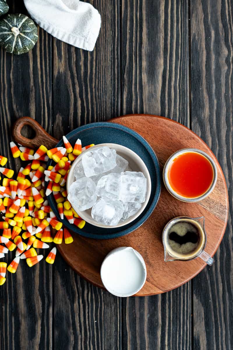 Ingredients used to make a candy corn latte sit on a wooden tabletop. Ingredients include candy corn, espresso, milk, and candy corn simple syrup.