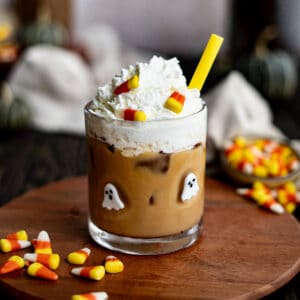 Feature image of a candy corn latte. A ghost glass is filled with a candy corn latte and sits on a countertop surrounded by candy corn.