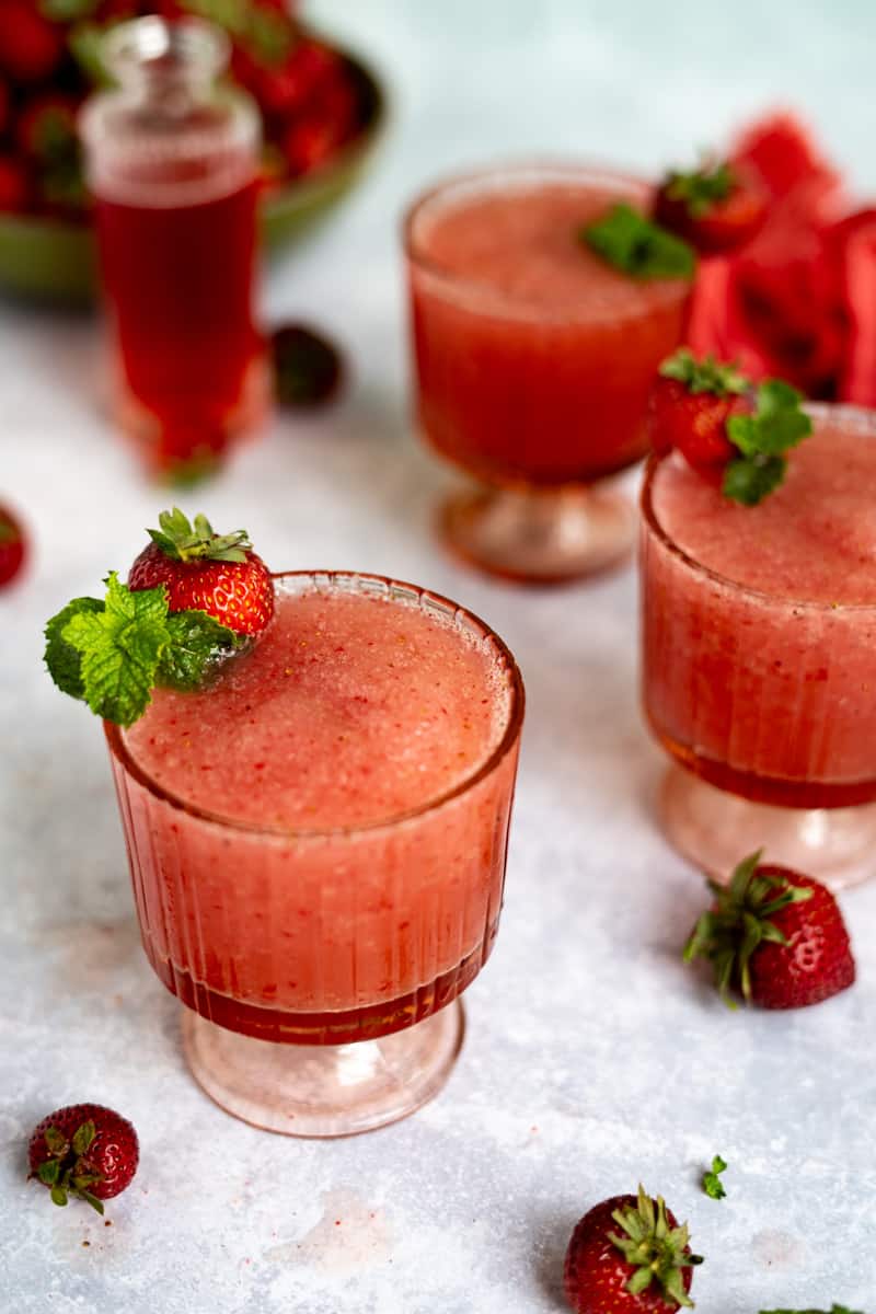 Hero image of frozé recipe. Three glasses filled with frozé are garnished with strawberries and mint, sitting on a grey countertop with berries strewn across it. A bottle of strawberry simple syrup sits in the background.