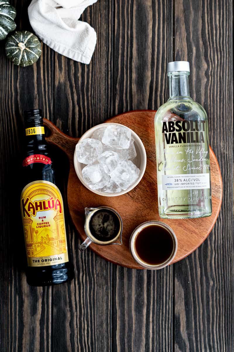 Ingredients used to make pumpkin espresso martinis sit on a wooden tabletop. The ingredients include vanilla vodka, Kahlua liqueur, espresso, pumpkin spice simple syrup, and ice.