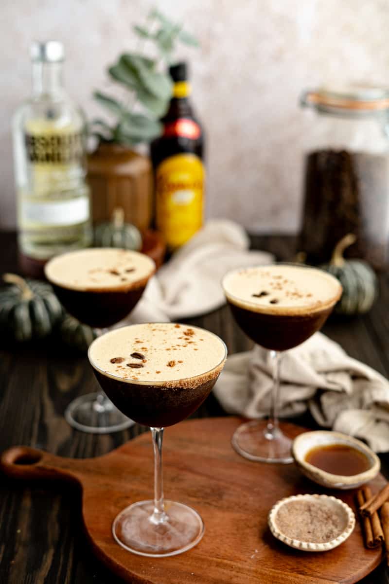 Hero image for a pumpkin espresso martini recipe. Three cocktail coupe glasses are filled with pumpkin espresso martinis. They sit on a wooden countertop that contains ingredients used to make the recipe.