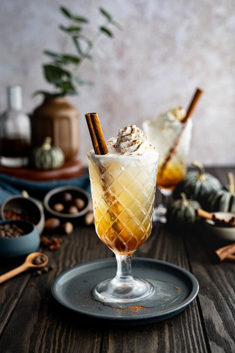 A pumpkin spice soda sits on a small blue dish on a dark wooden countertop. The soda is garnished with whipped cream, pumpkin spice mix, and a cinnamon stick.