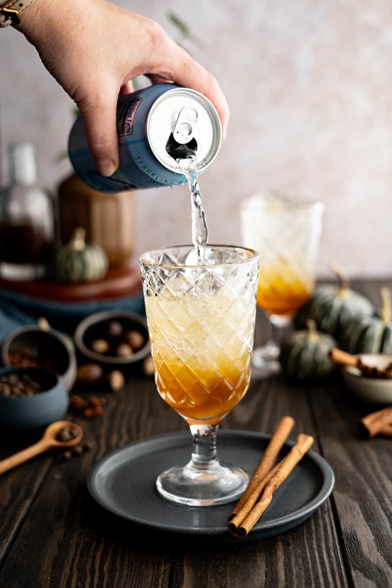 A hand from out of frame pours soda water into a glass with pumpkin spice simple syrup.