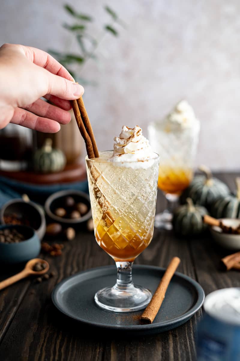 A hand from out of frame garnishes a glass of pumpkin spice soda with a cinnamon stick.