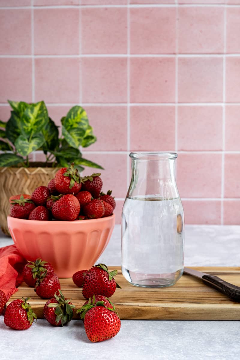A bowl of strawberries and a small bottle of water sit on a cutting board in preparation of making strawberry ice cubes.