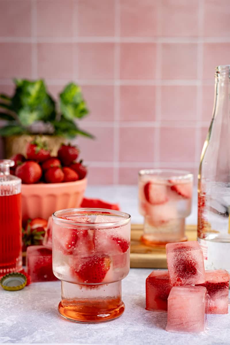 Hero image of strawberry ice cubes. A small pile of cubes sits to the right of a glass of homemade strawberry soda, filled with the ice cubes.
