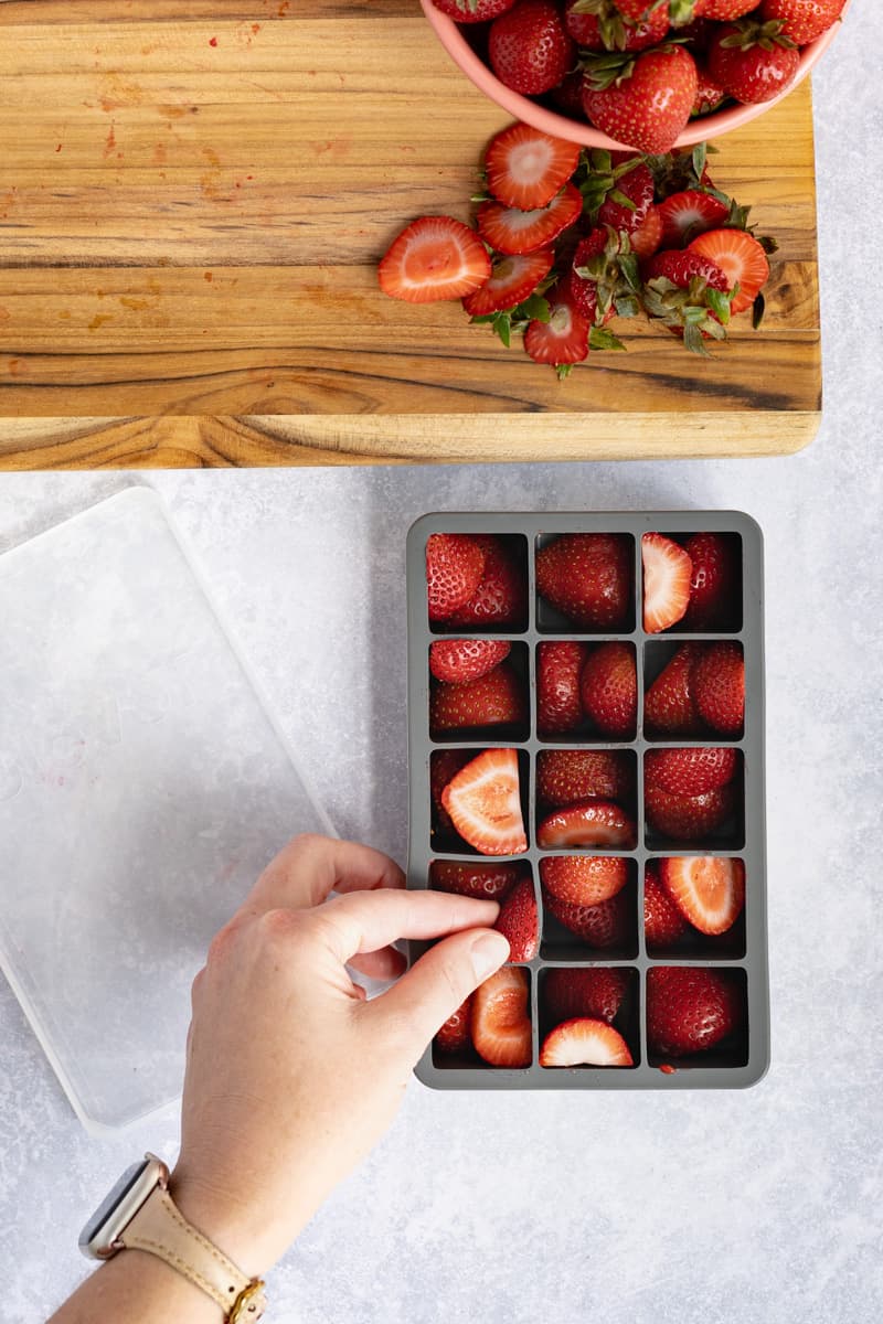 A hand from out of frame pushes strawberries into the sides and bottom of an ice cube tray.