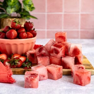 Feature image of strawberry ice cubes. Frozen strawberry ice cubes sit on a grey countertop with a pink tile background. The ice cubes are piled up, and you can see pieces of strawberries peeking through the ice cubes.A bowl of strawberries sits to the left of the pile of ice.