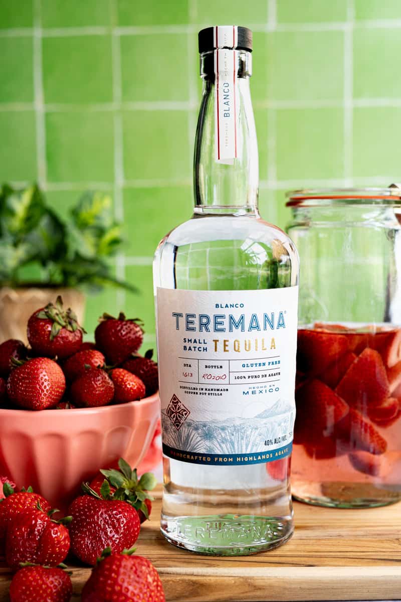 A bottle of tequila sits next to a bowl of strawberries on a wooden cutting board. A jar of strawberry infused tequila sits in the background.