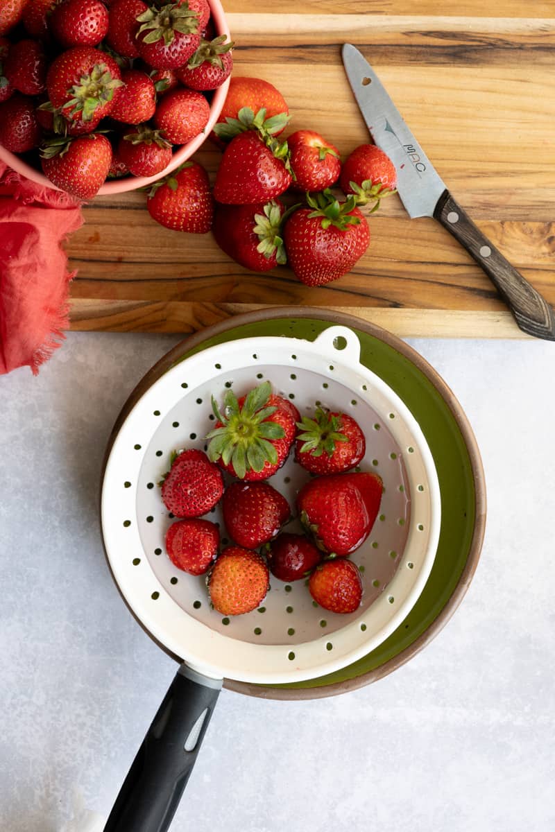 A small colander is filled with strawberries in a bowl of water in preparation of making strawberry infused tequila.