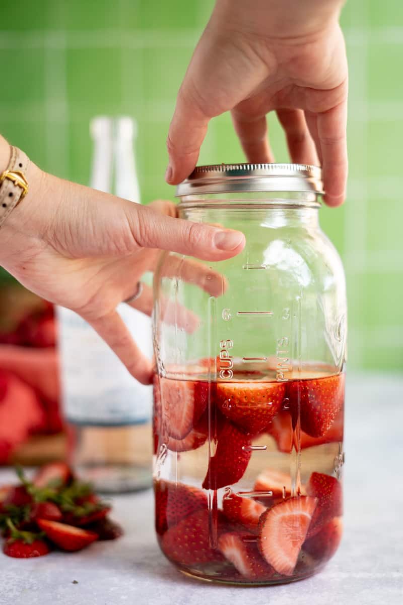 Hands from out of frame seal a large mason jar filled with strawberry infused tequila.