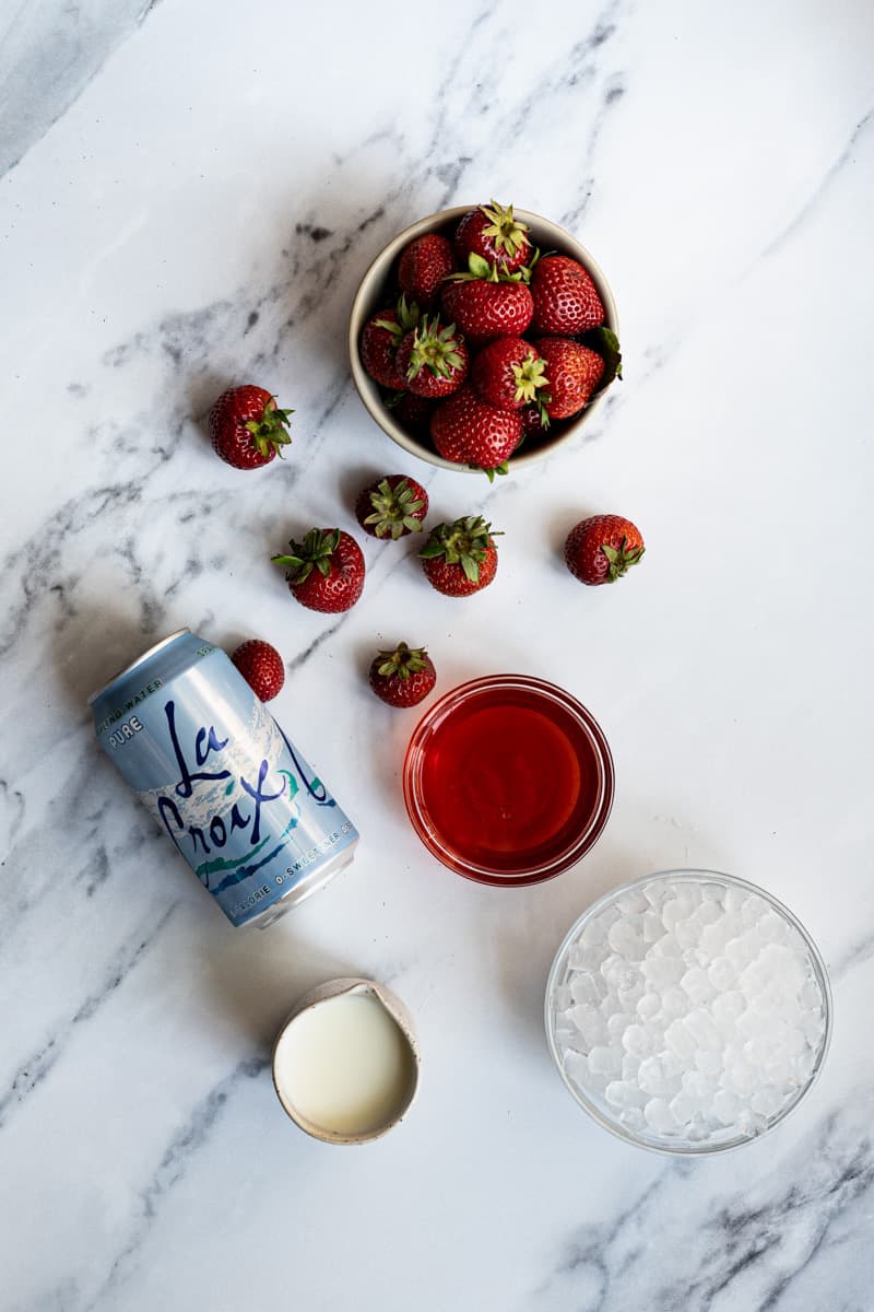 ingredients used to make a strawberry Italian soda. The ingredients are laid out on a marble countertop.