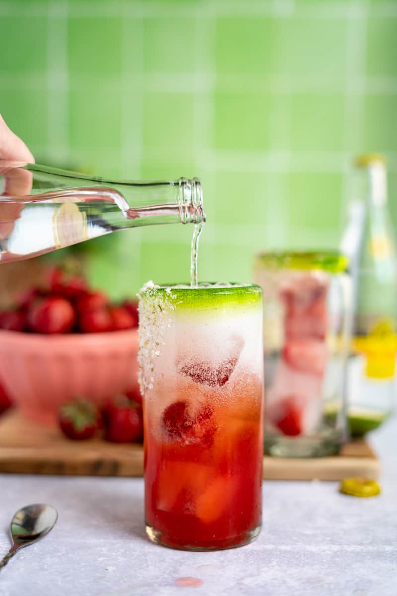 A bottle of topo chico is being poured into a cocktail glass to finish making strawberry ranch water.