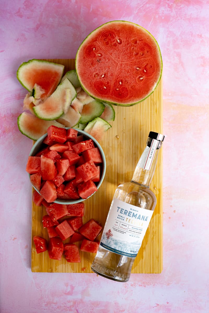 Ingredients used to make watermelon infused tequila. There is a bowl of watermelon chunks and a bottle of tequila on a cutting board.