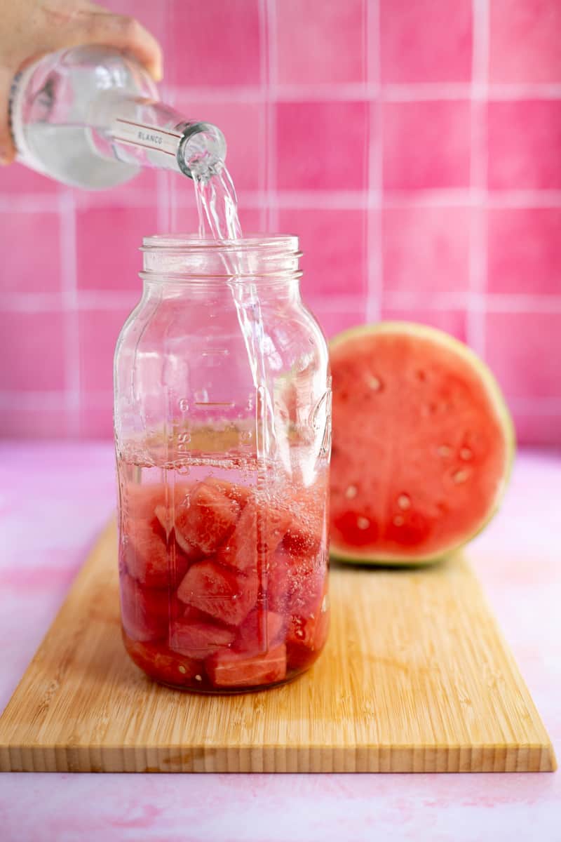 Pouring tequila into the large glass mason jar filled with cubes of watermelon.
