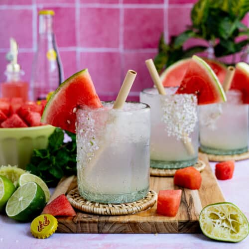 Feature Image for watermelon ranch water. Three glasses of watermelon ranch water sit on a countertop surrounded by ingredients used to make the cocktail.