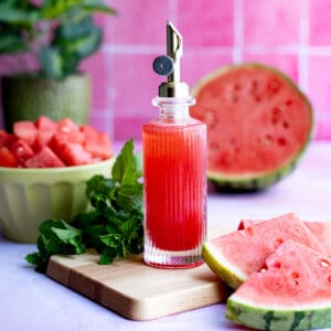 Hero image of watermelon simple syrup. A bottle of watermelon simple syrup sits on a small wooden cutting board.