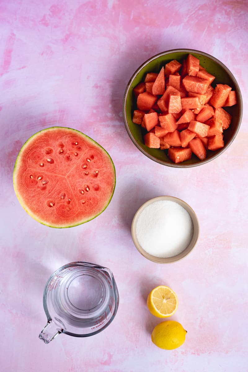 Ingredients used to make watermelon simple syrup sit on a pink countertop.