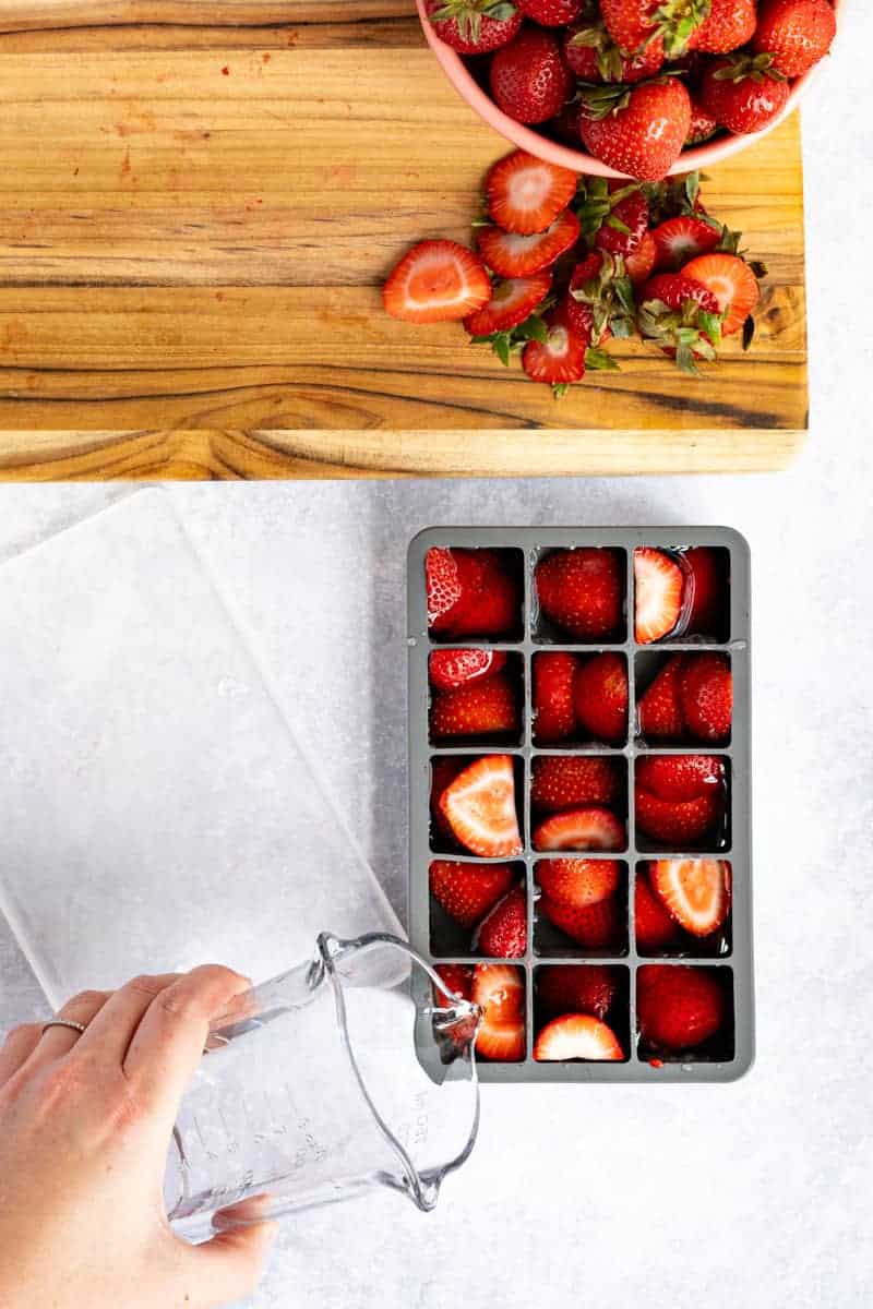 A hand from out of frame pours water overtop strawberries in an ice cube tray.