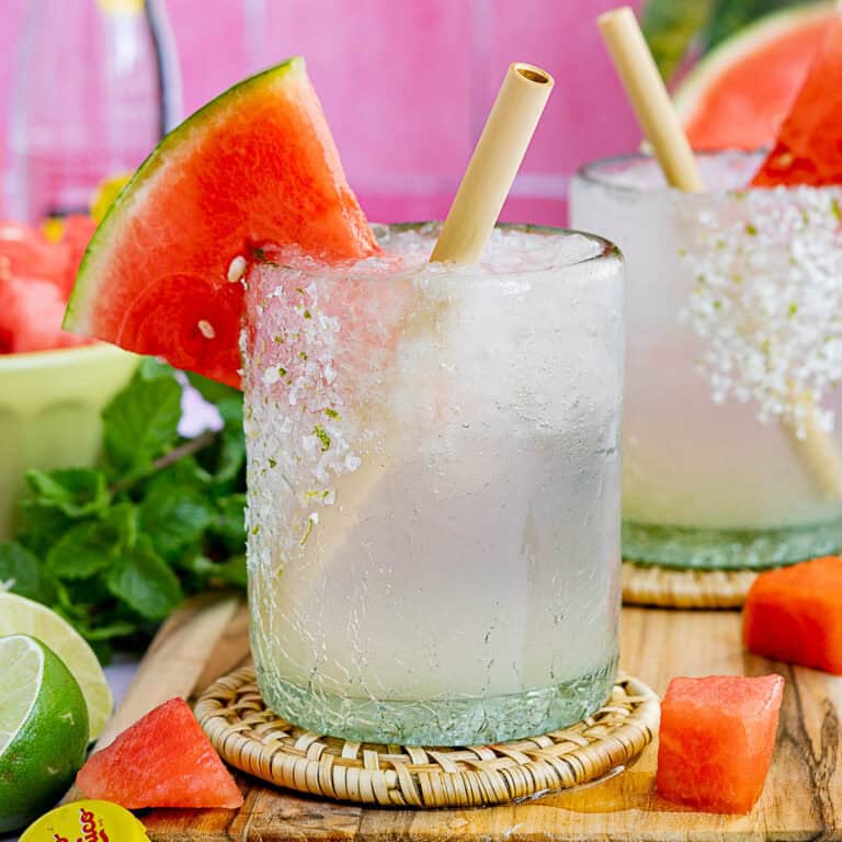 A glass of clear, icy watermelon ranch water with a salted rim, garnished with a watermelon wedge and straw, sits on a coaster surrounded by lime, mint, and watermelon pieces. Another similar drink is in the background.
