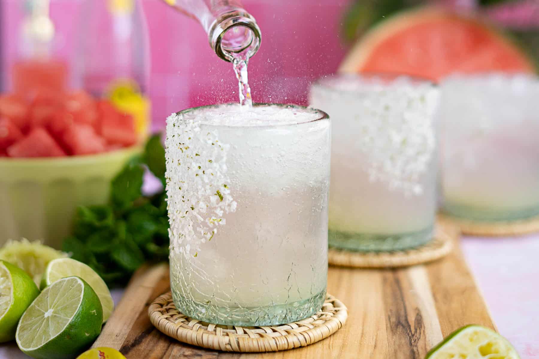 A glass with a salted rim is being filled with a clear, fizzy drink—perfect for making refreshing watermelon ranch water. Sliced limes, mint leaves, and a bowl of watermelon rest on a wooden board, with another glass in the background.