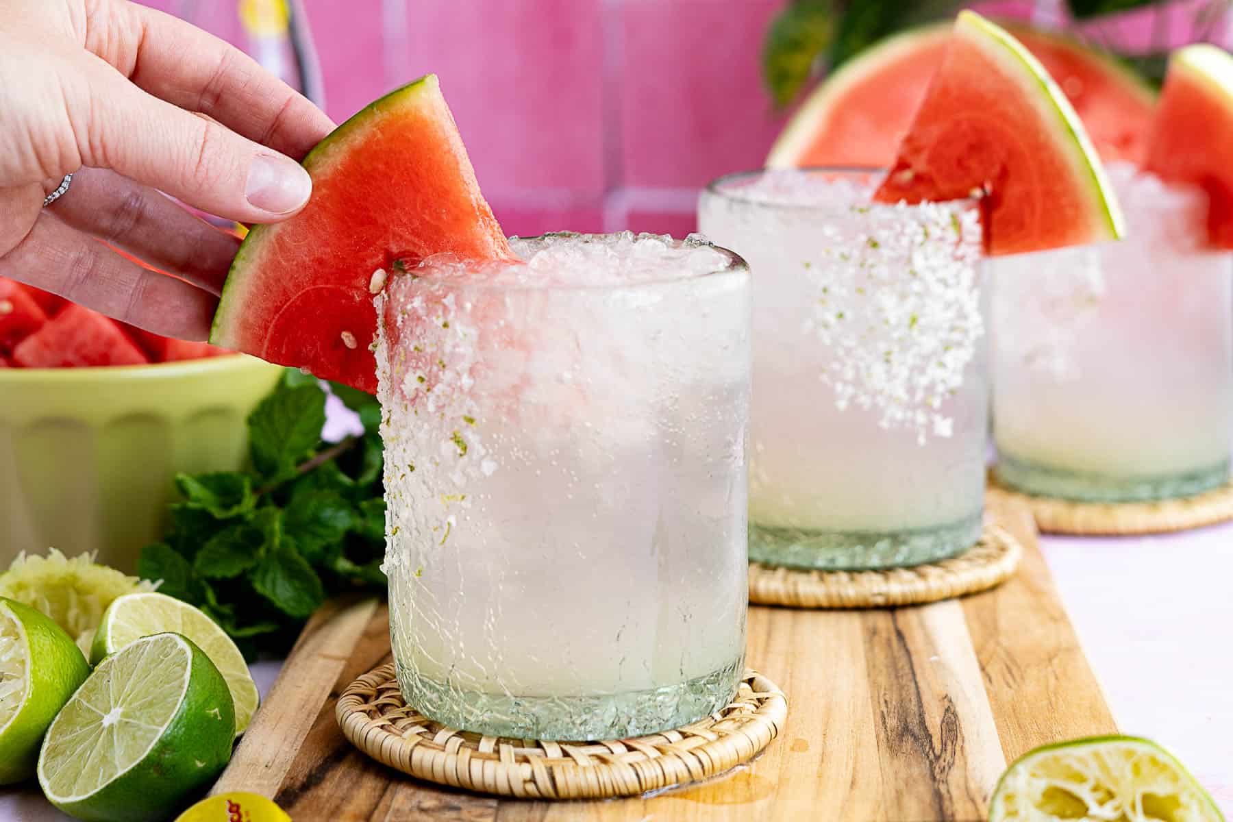 A hand places a slice of watermelon on the rim of a glass filled with icy watermelon ranch water. The salted-rim glass is garnished with watermelon, while more drinks, lime, and mint rest on a wooden board in the background.