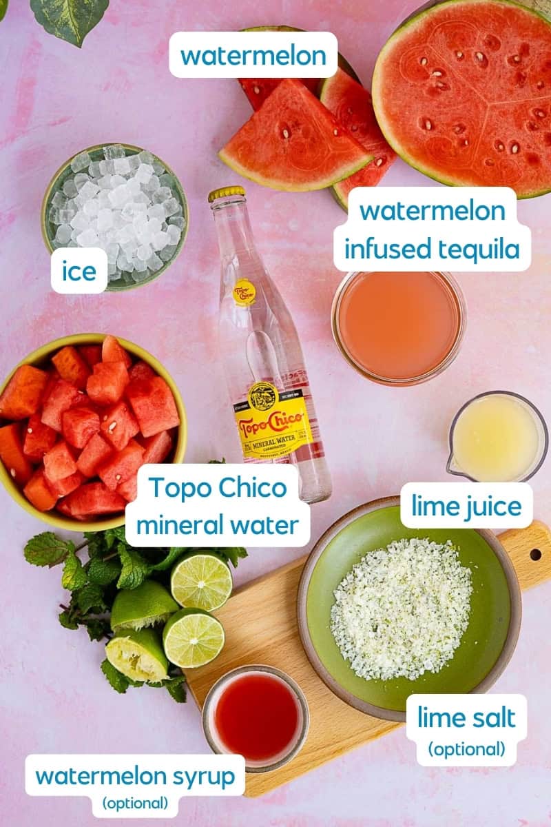 Ingredients for a refreshing watermelon ranch water cocktail are displayed: watermelon slices, diced watermelon, ice, watermelon-infused tequila, lime juice, Topo Chico mineral water, lime salt, and watermelon syrup.