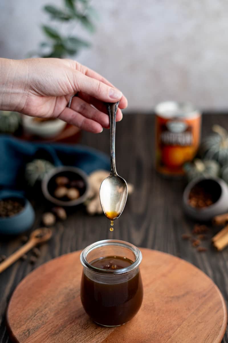 A hand from out of frame drips pumpkin spice simple syrup from a spoon into a small jar on a wooden cutting board.