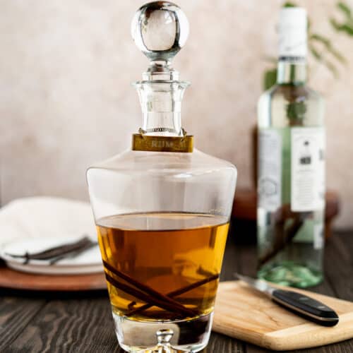 Feature image of a bottle of vanilla infused rum sits on a wooden table top. A bottle of rum being infused sits in the background, and a plate of vanilla beans is in the background as well.
