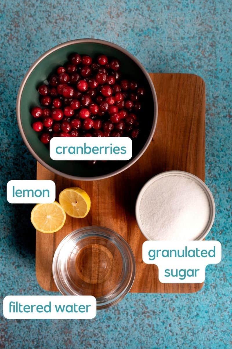 Ingredients used to make a homemade cranberry simple syrup sit on a wooden cutting board against a teal countertop. The ingredients include fresh cranberries, granualted sugar, filtered water, and lemons.