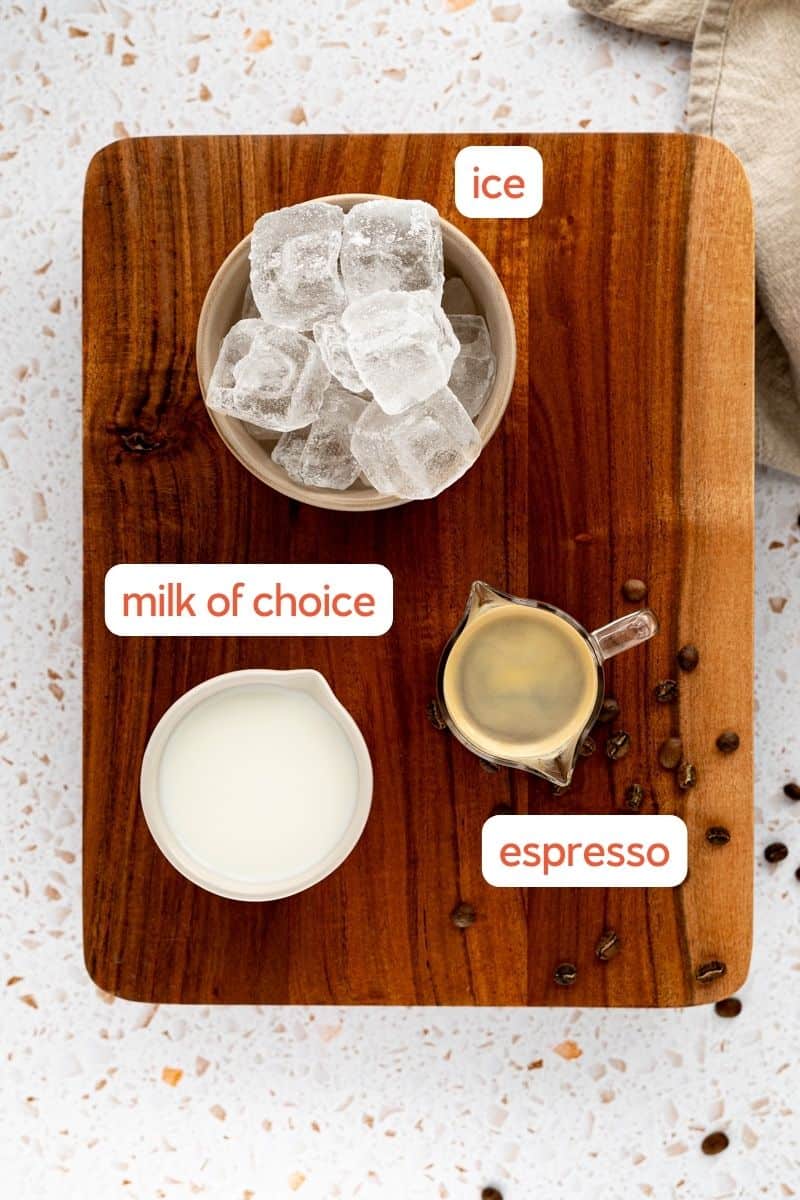 ingredients used to make an iced cortado beverage sit on a wooden cutting board. The photo is labeled with the ingredients: espresso, ice, and milk. There are espresso beans scattered on the countertop as well as a linen napkin.