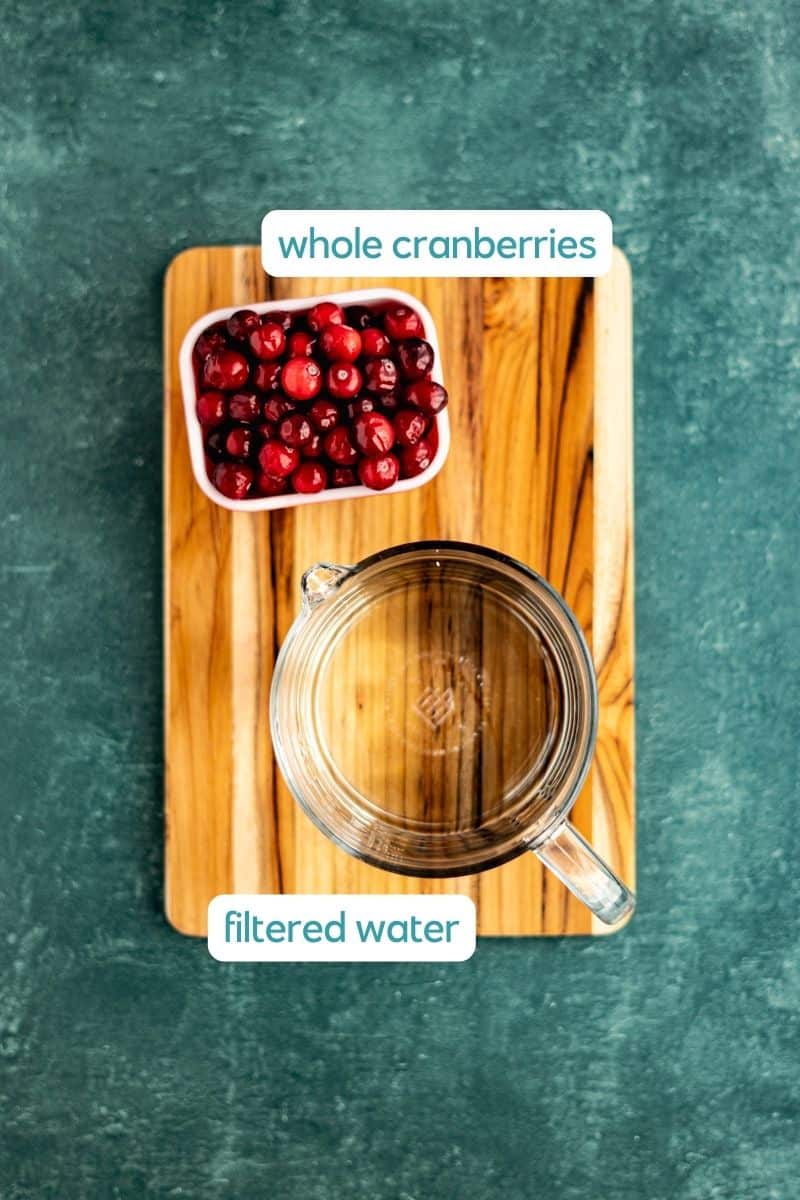 Ingredients used to make cranberry ice cubes sit on a small wooden cutting board on a green countertop.