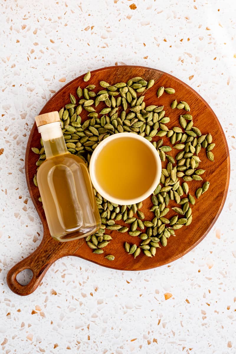 A bottle of cardamom simple syrup sits on a round wooden cutting board of cardamom pods. A small bowl of simple syrup sits in the middle of the cutting board.