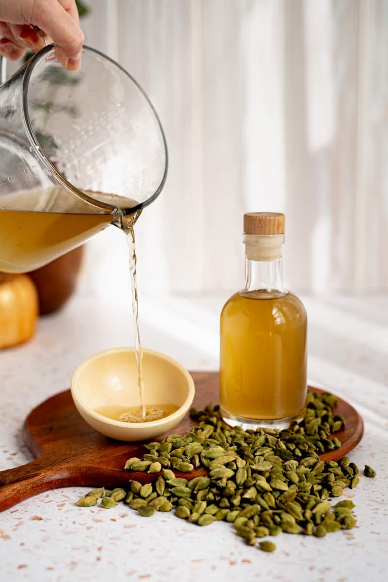 A measuring glass slowly pours cardamom simple syrup into a bowl.