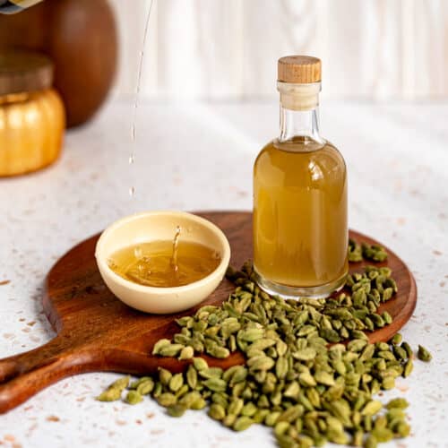 A small bowl of cardamom simple syrup sits next to a bottle of the syrup on a round wooden cutting board.