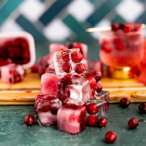 A small pile of cranberry ice cubes sits on a green countertop with a small wooden cutting board in the background.