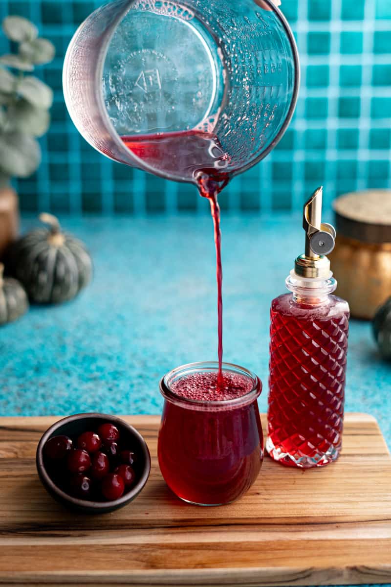 Pouring cranberry simple syrup from a measuring glass into a small glass jar to show the viscosity of the simple syrup.