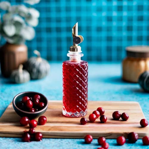 A bottle of cranberry simple syrup sits on a wooden cutting board in a teal kitchen. A small bowl of cranberries are scattered on the cutting board.