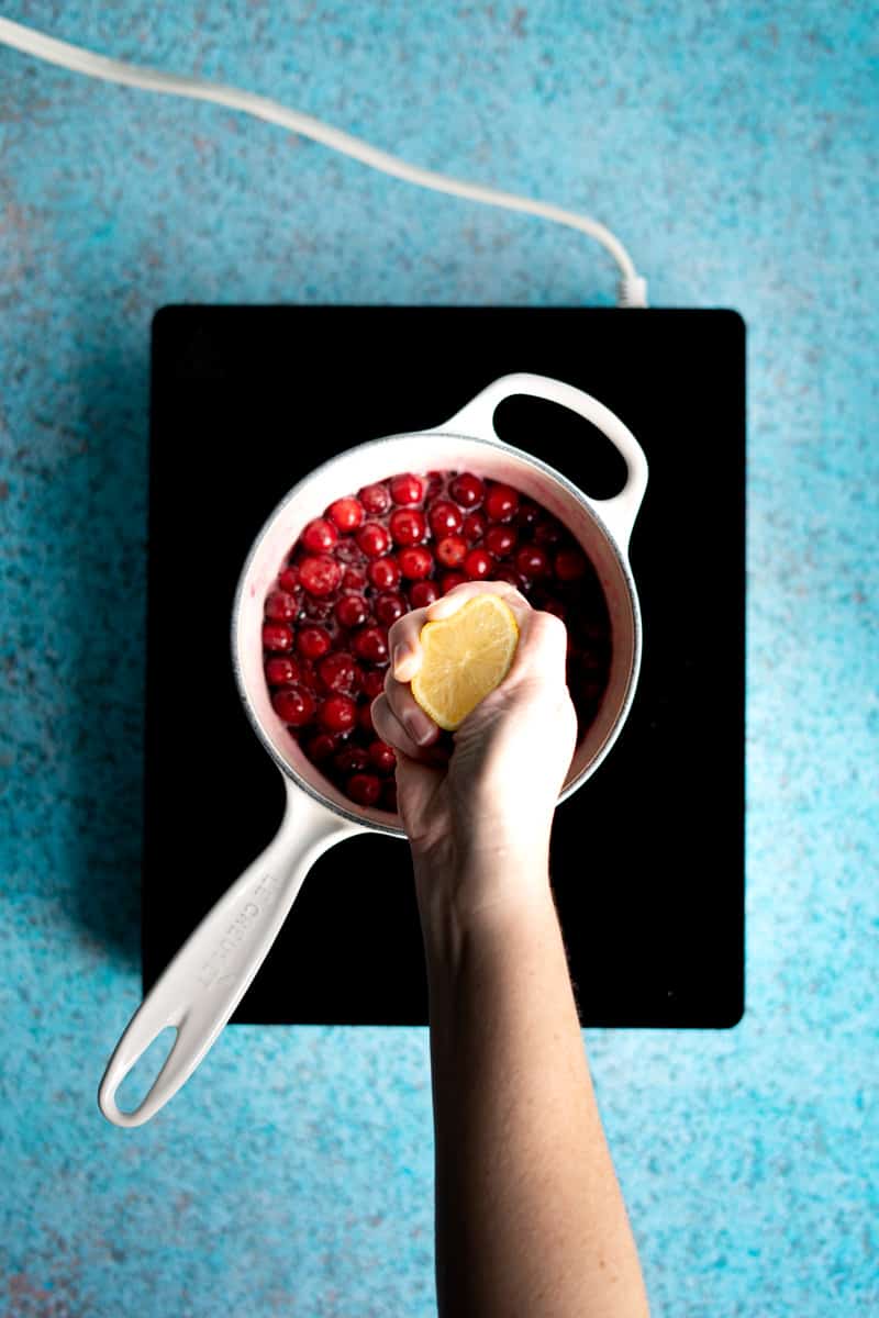 A hand from out of frame squeezes a lemon into the small saucepan to add lemon juice to the cranberry simple syrup.