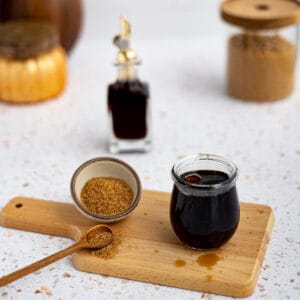 A small glass jar of demerara simple syrup sits on a wooden cutting board next to a bowl of demerara sugar.
