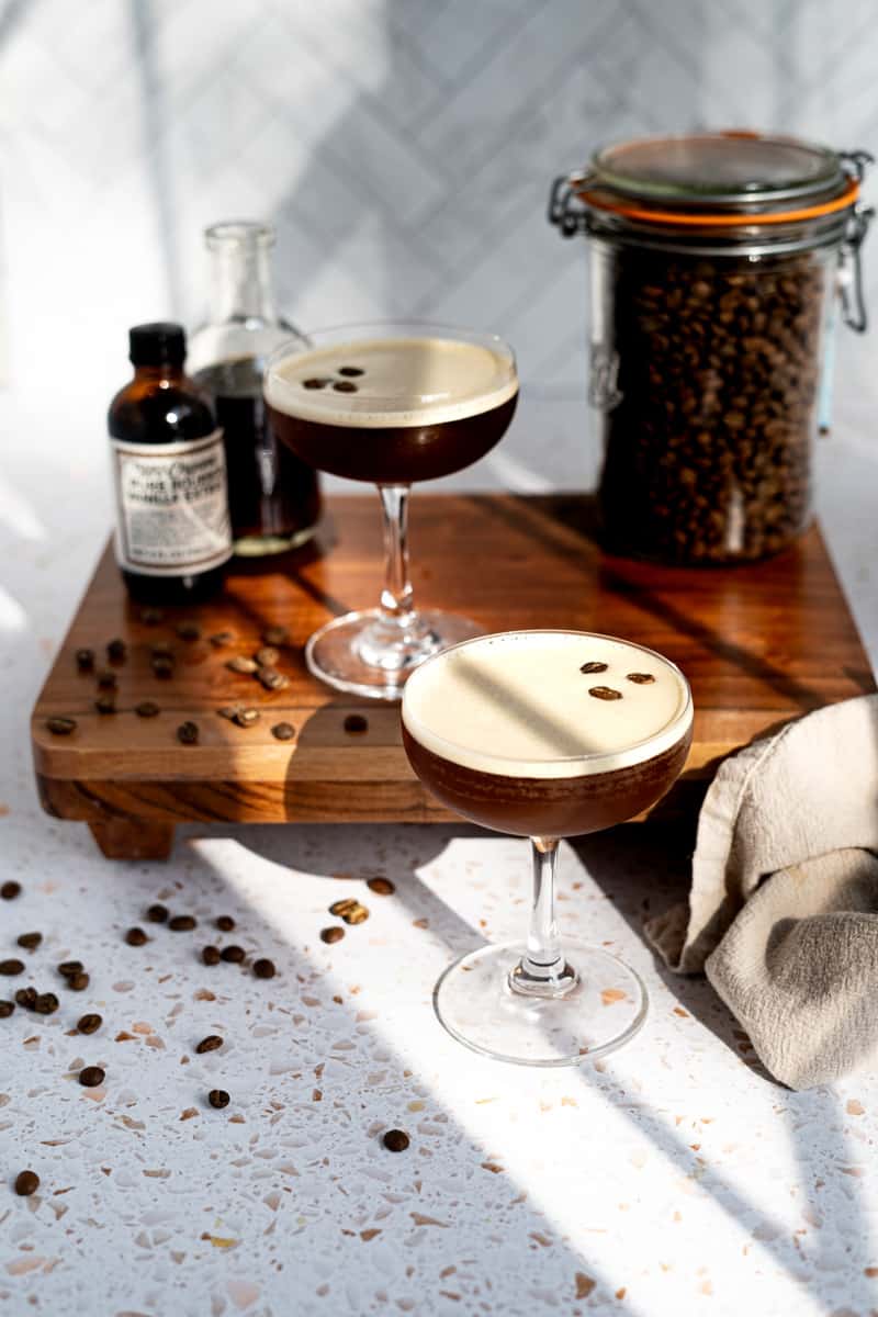Feature image for an espresso martini mocktail. Two glasses of virgin espresso martinis sit on a countertop next to the ingredients used to make them.