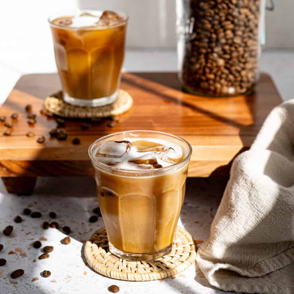 Two iced cortados sit on a countertop. There is a jar of coffee beans in the background, and coffee beans are scattered on the countertop.